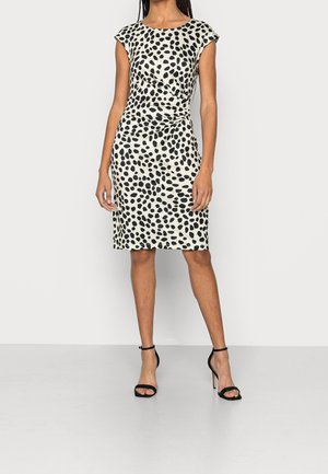 Vrouw draagt een knielange, witte jurk met zwarte onregelmatige stippen, gerimpelde taille, korte mouwen en zwarte sandalen met hoge hakken.