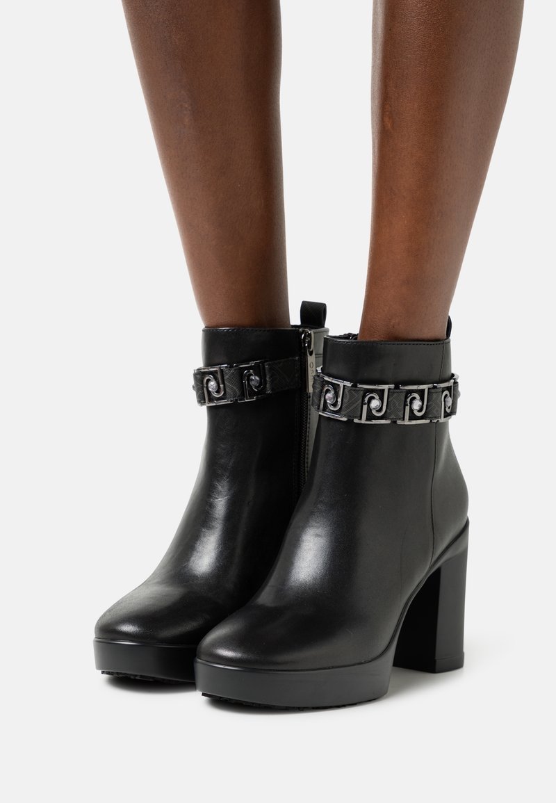Bottines noires en cuir avec talons blocs et détail de chaîne métallique décorative portées sur des jambes nues sur fond blanc.