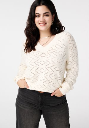 Roomkleurige, gebreide trui met V-hals, voorzien van een zigzagpatroon en opengeweven structuur. Gecombineerd met donkere jeans voor een casual uitstraling.