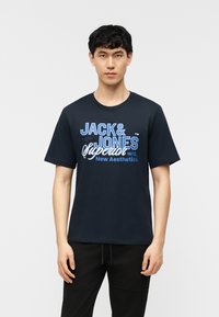 Jeune homme debout, face à l'avant, portant un t-shirt noir "Jack & Jones Superior New Aesthetics" et un pantalon noir, sur un fond uni.