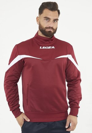 Mężczyzna z brodą w bordowej sportowej bluzie LEGEA z białymi paskami na rękawach, stoi z rękami w kieszeniach na białym tle.
