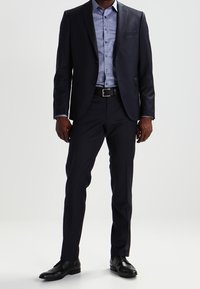 Marineblauw pak met een slank ontwerp, gecombineerd met een lichtblauw geruit overhemd. Gladde stof, enkele knoopsluiting en zwarte leren schoenen.