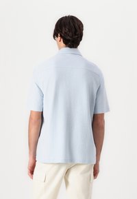 Camisa de manga corta azul claro hecha de una tela texturizada, con cuello y un dobladillo recto. La parte posterior es lisa con sutiles detalles de costura.