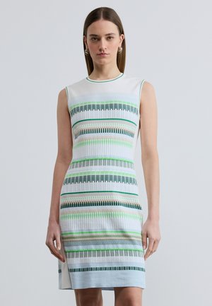 Sieviete valkā bezroku ceļgalu garuma kleitu ar horizontālām zaļām, zilām, bēšām un melnām svītrām, stāvot pret vienkāršu gaišu fonu.