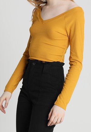 Persoon draagt een mosterdgele geribde top met lange mouwen en V-hals en een zwarte broek met hoge taille tegen een effen achtergrond.