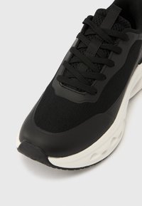 Μαύρο αθλητικό παπούτσι με επάνω μέρος από πλέγμα και συνθετικό υλικό, μαύρα επίπεδα κορδόνια και λευκή σόλα με υφή και στρογγυλεμένη μύτη.