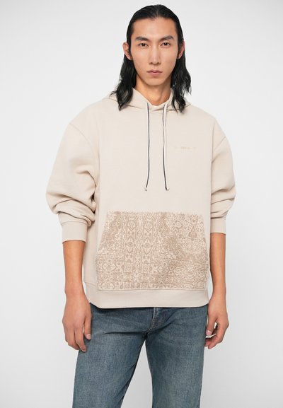 Bežs džemperis ar teksturētu rakstu priekšējo kabatu, ar aukliņu kapuci un ribota malām. Virs kabatas ir diskrēts logo.