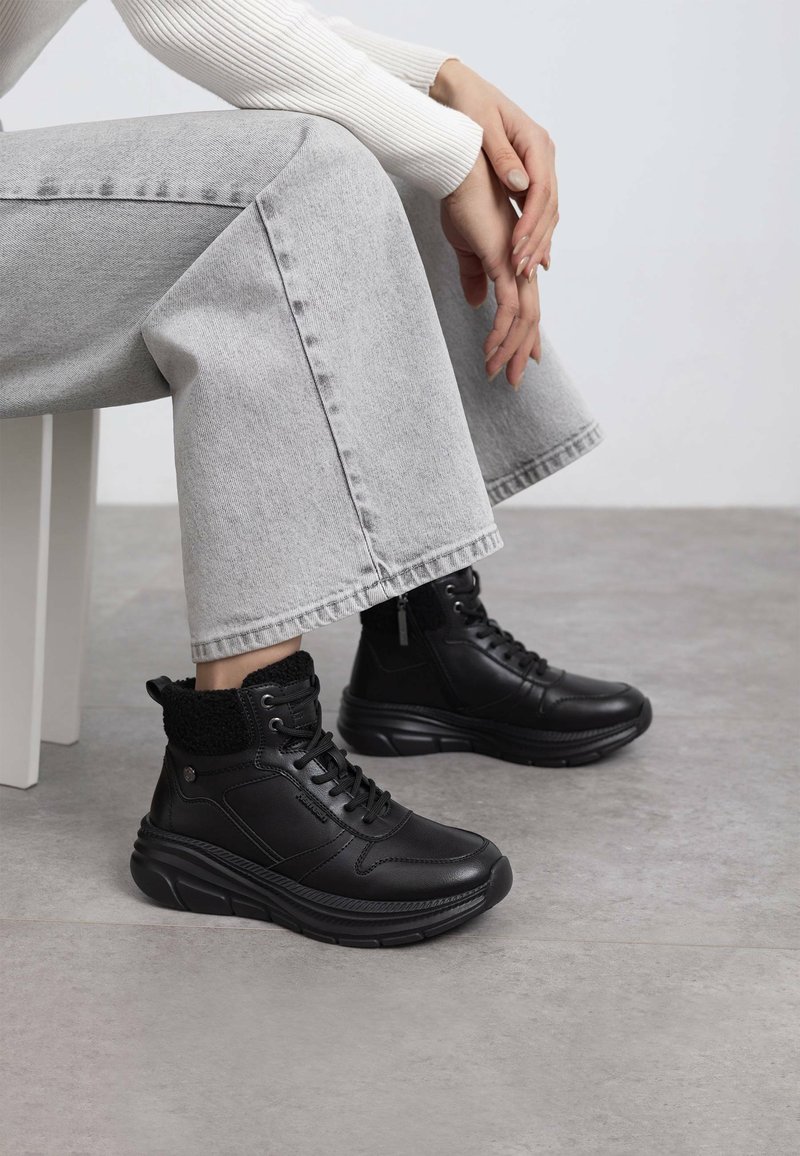 Zapatos altos de cuero negro con un cuello de punto negro texturizado, parte delantera con cordones y suela gruesa, combinados con jeans anchos de color gris claro.