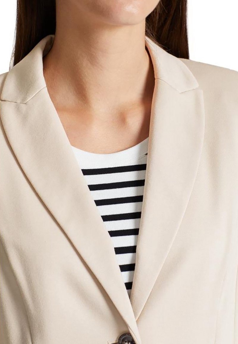 Femme portant un blazer beige sur une chemise blanche et noire à rayures horizontales, visible du menton jusqu'au milieu de la poitrine.