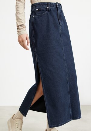 Donkerblauwe lange denim rok met zijschuif gedragen met een beige top met lange mouwen, beige sokken en beige sneakers op een lichte achtergrond.