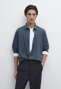 Camicia grigia con sottocamicia bianca; vestibilità comoda, maniche lunghe arrotolate ai polsini, abbinata a pantaloni scuri, texture liscia.