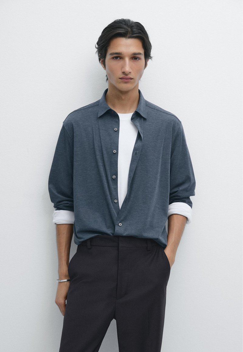 Camicia grigia con sottocamicia bianca; vestibilità comoda, maniche lunghe arrotolate ai polsini, abbinata a pantaloni scuri, texture liscia.