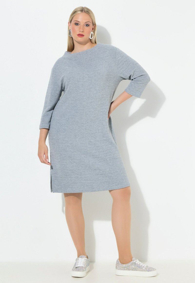 Šedé pletené šaty s volným střihem, tříčtvrtečními rukávy, bočními rozparky a kulatým výstřihem. Doplněné třpytivými stříbrnými teniskami.