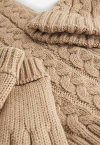 Beige kabelbrei trui met dikke geribbelde manchetten en een gestructureerd patroon van met elkaar verweven kabels, wat de zachtheid en warmte benadrukt.