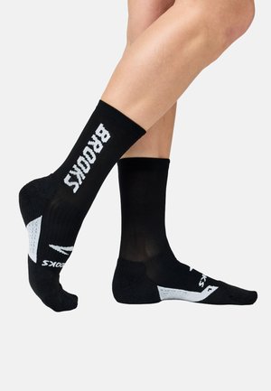 Noge v črnih nogavicah do polovice meč z belimi poudarki in napisom "BROOKS" na strani na belem ozadju.