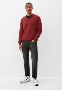 Rode v-hals trui met lange mouwen, gedragen over een zwart T-shirt. Gecombineerd met donkere jeans en witte sneakers met zwarte accenten. Strak ontwerp.