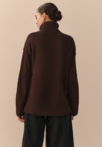 Brauner übergroßer Pullover mit hohem, geripptem Kragen, überschnittenen Schultern und geripptem Saum. Weicher Stoff; getragen mit weit geschnittenen schwarzen Hosen.
