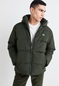 Giacca a vento imbottita verde scuro con zip frontale, cappuccio e tasche anteriori. Presenta un design trapuntato e un piccolo logo sul petto.
