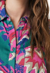 Chemise boutonnée colorée présentant un mélange de motifs floraux roses, verts et bleus. Texture douce avec une poche poitrine et des boutons ronds.
