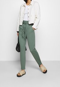 Woman wearing a white cropped jacket, white top, green drawstring pants, beige slide sandals, holding a black handbag.