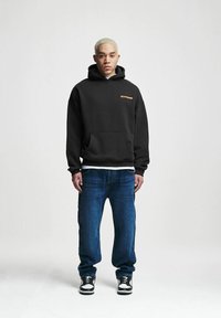 Black oversized hoodie with kangaroo pocket, orange logo on chest, paired with blue jeans and black-and-white sneakers. Plain background.