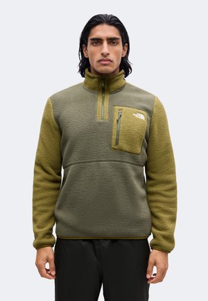 Hombre con cabello oscuro de longitud media que lleva un jersey polar verde y oliva con textura, con un bolsillo en el pecho con cremallera y pantalones negros.