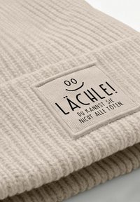 Beige knitted fabric with a sewn white tag showing a smiley face and German text "Lächle! Du kannst sie nicht alle töten."