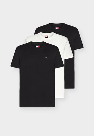 Homme portant un t-shirt blanc, un jean bleu ample, des baskets blanches, et tenant un sac en cuir bleu marine avec un bracelet en or au poignet.