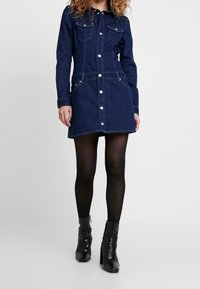 Tumapaita denim tummansininen nappikiinnityksellä, rintataskuilla ja sivutaskuilla. Omistaa kapean siluetin ja polven puoliväliin ulottuvan pituuden.