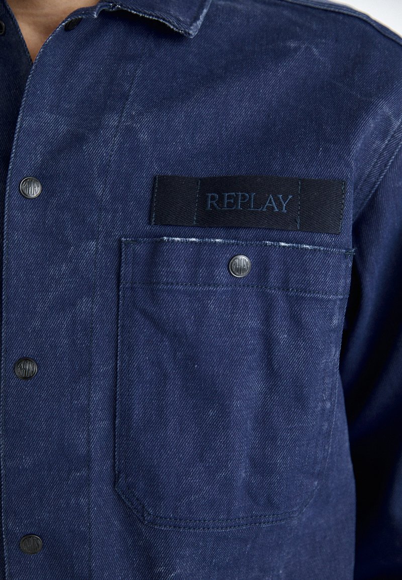 Granatowa koszula dżinsowa z metalowymi zatrzaskami, z przednią kieszenią z czarną metką z napisem "REPLAY" w niebieskim kolorze.