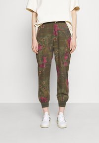 Groene cargobroek met bloemenprint en roze accenten, voorzien van zijzakken, elastische boorden en een relaxte pasvorm. Ggedragen met witte sneakers.