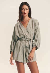 Wikkelromper in lichtgroene stof met zwarte polka dots, met pofmouwen, een getailleerde taille met strik en een V-hals.