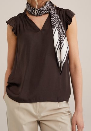 Vrouw draagt een zwarte mouwloze blouse, crèmekleurige broek en een zwart-wit sjaaltje om haar nek geknoopt.