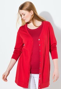 Cardigan rosso con maniche lunghe, caratterizzato da una chiusura a bottoni e un orlo a costine. Indossato sopra un top rosso a maniche corte, con pantaloni bianchi.