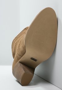 Stivale con tacco in suede color beige, con superiore testurizzato, perforazioni e tacco in legno marrone. La suola è di un colore chiaro con un motivo a righe.