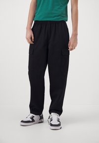 Musta työhaalari, jossa on joustava vyötärönauha ja kaksi sivutaskua, yhdistettynä valkoisiin lenkkareihin mustilla yksityiskohdilla ja vihreään t-paitaan.