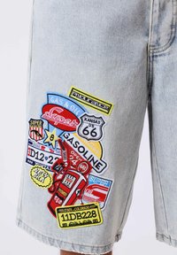 Pantalones cortos de mezclilla clara con parches temáticos de gasolineras coloridos que incluyen la Ruta 66, surtidores de gasolina y matrículas vintage en el muslo.