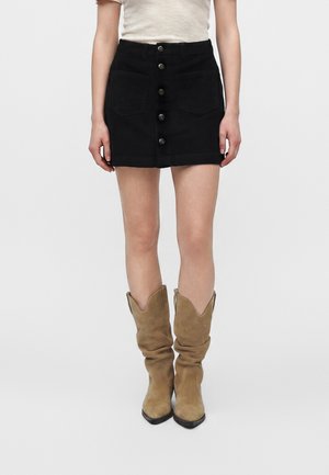 Kvinde iført sort knap-front nederdel, beige ribbet top og brune ruskindsløse støvler til midt på læggen, stående mod en hvid baggrund.