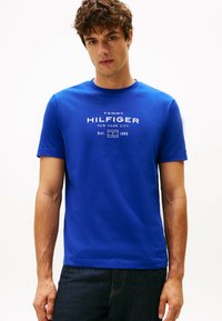 Blå bomulds T-shirt med rund hals, med et hvidt logo print. Korte ærmer og lige kant. Bæres casual med mørke jeans.