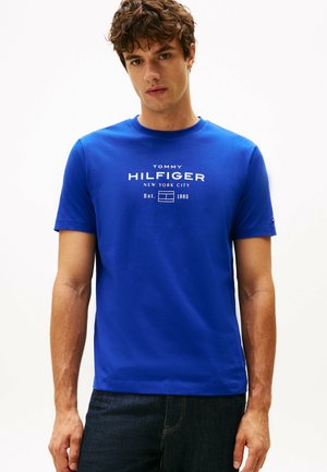 Camiseta de algodón azul con cuello redondo, que presenta un estampado de logo en blanco. Mangas cortas y dobladillo recto. Llevada de manera casual con jeans oscuros.