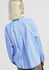 Camicia a maniche lunghe azzurro chiaro realizzata in tessuto traspirante, con dettagli plissettati sulla schiena, polsini con bottoni e una vestibilità rilassata.