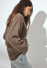 Bruine gebreide trui met een hoge ribbenkraag en geribde zoom, met een relaxte pasvorm en zachte textuur, gecombineerd met lichte spijkerbroek.