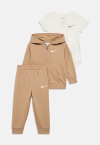 Detské oblečenie Nike pre batoľa zahŕňa béžovú mikinu na zips, zodpovedajúce béžové nohavice a krémové body, všetko s bielymi logami.
