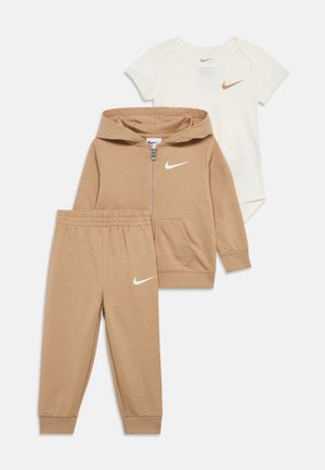 Nike-tøj til småbørn med en beige lynlåshættetrøje, matchende beige bukser og en cremefarvet heldragt, alle med logoaccents i hvid.