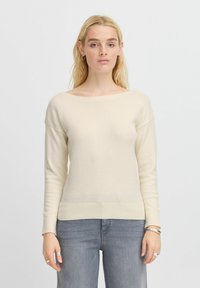 Maglione color crema con ampia scollatura a barca, maniche lunghe, polsini e orlo a coste. Tessuto morbido, abbinato a jeans grigi chiari.