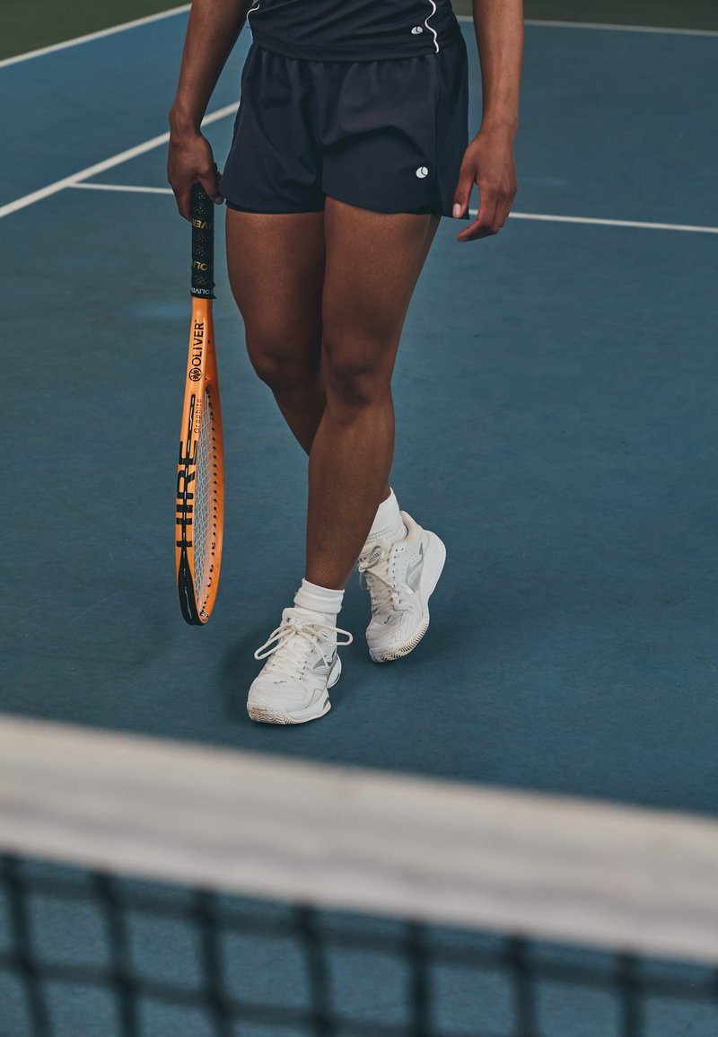 Raqueta de tenis con marco naranja y empuñadura negra sostenida por una persona que lleva pantalones cortos negros y zapatillas blancas con suelas texturizadas, en una cancha azul.