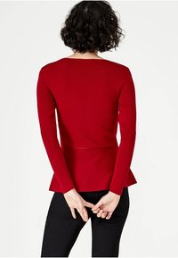 Czerwony sweter z długim rękawem, faktura w prążki, dopasowana góra, peplum na dole, widok z tyłu, w połączeniu z czarnymi spodniami, dłonie złożone z tyłu.