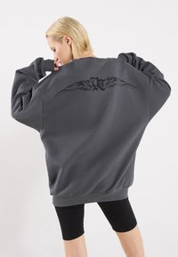 Felpa oversize grigia scura con maniche lunghe, caratterizzata da un design di ali nere sulla schiena. Texture liscia e polsini a coste.