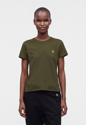 Femeie cu păr scurt, purtând un tricou verde măsliniu și pantaloni negri, ambele cu mici sigle albe cu jucător de polo pe partea stângă.