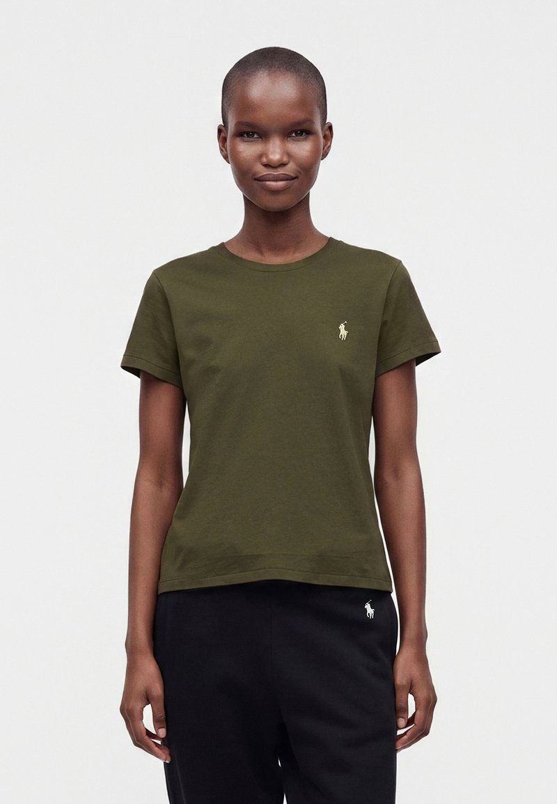 Femme aux cheveux courts portant un t-shirt vert olive et un pantalon noir, tous deux avec de petits logos blancs de joueur de polo sur le côté gauche.